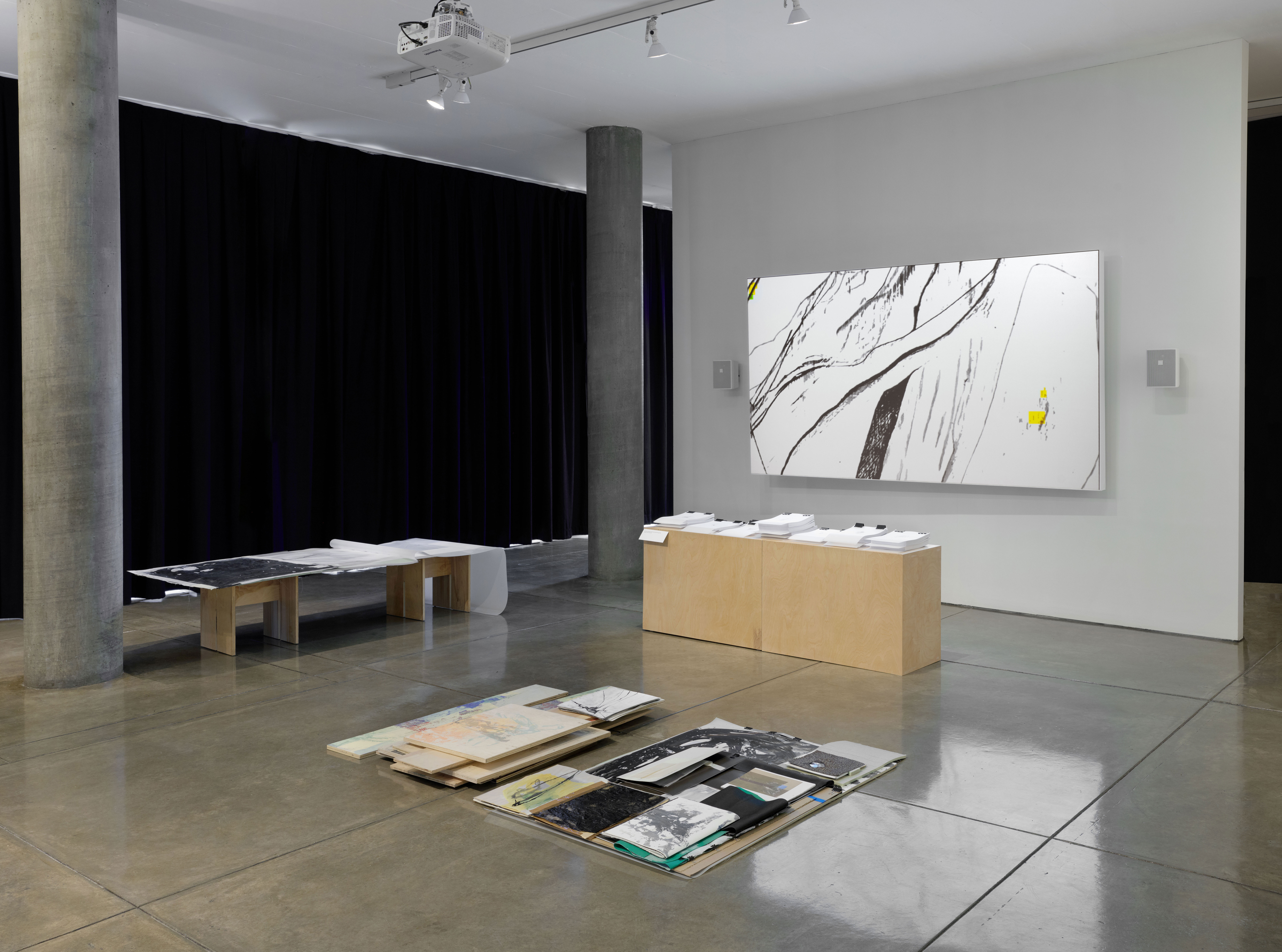 Photograph of an art installation by artist Caroline Coolidge with a projected screen in the back with abstract black, white, and yellow lines forming the background, and mixed media artworks across two tables and the concrete floor.