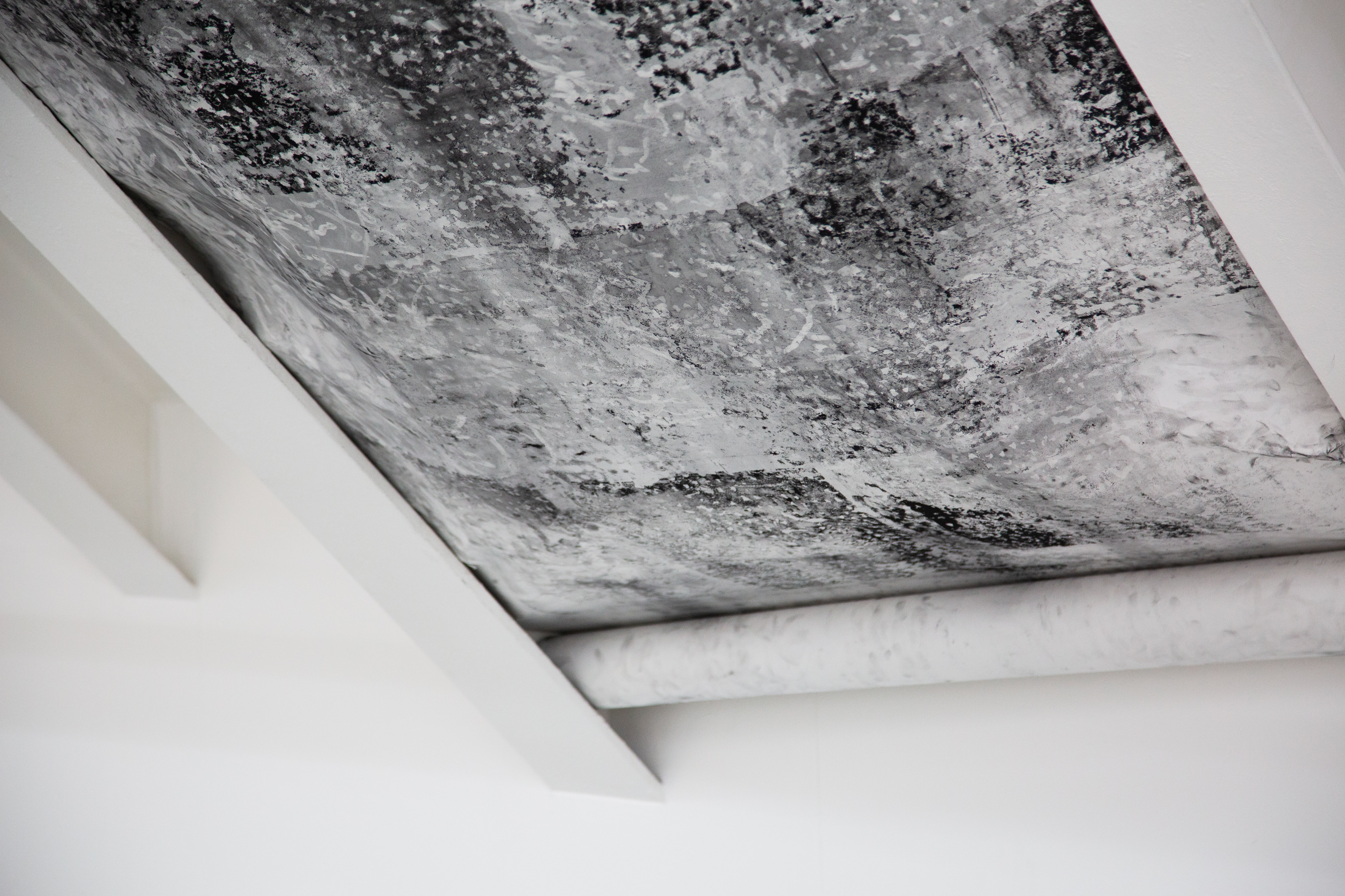 A photograph showing an angled view of the lower half of a black and white, abstract, highly textural, charcoal drawing, installed between two white slanted metal beams and rolled up at the bottom.