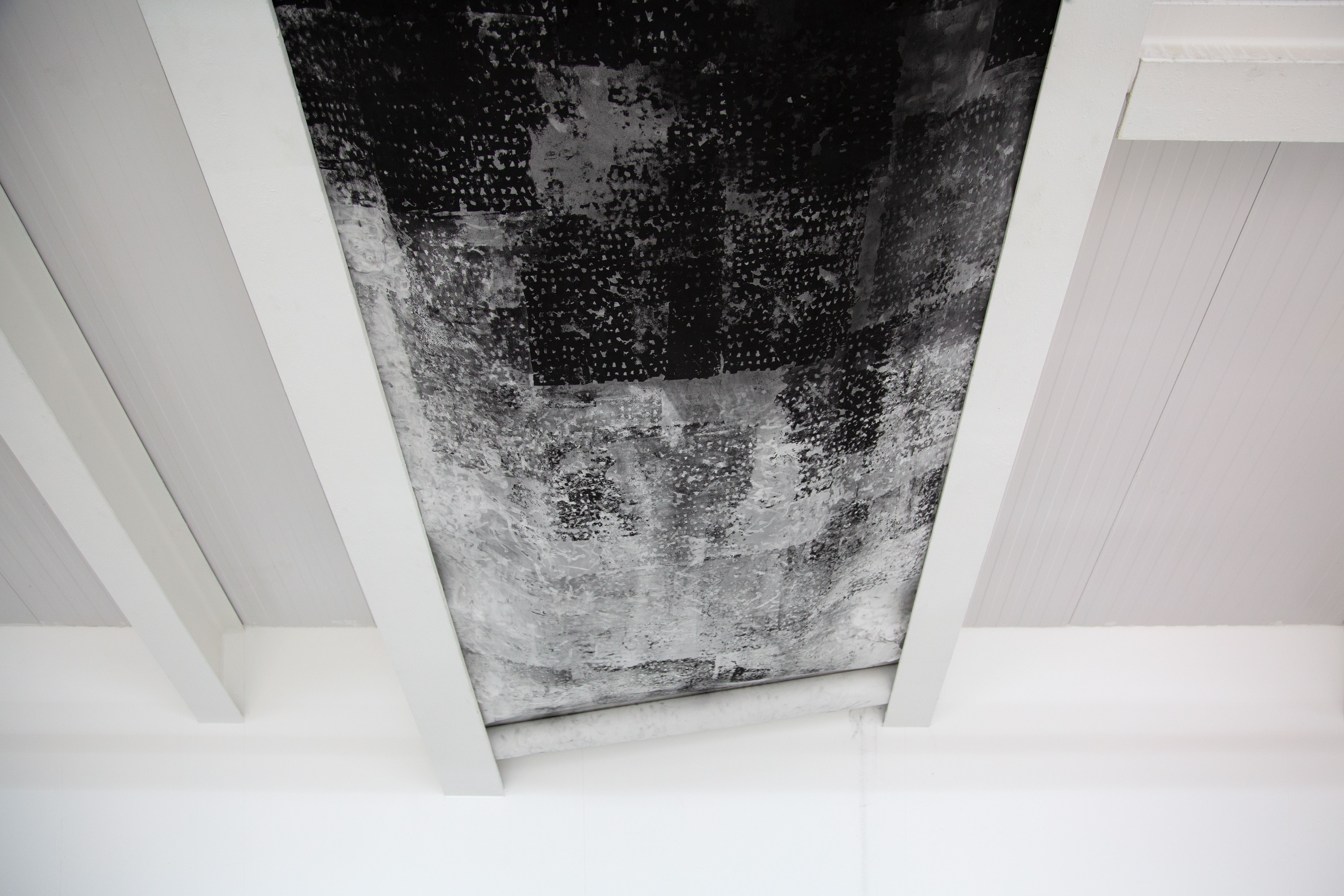 A photograph showing a black and white, abstract, highly textural, charcoal drawing, installed between two white slanted metal beams and rolled up at the bottom, seen from a font view, with the top of the drawing cut off.