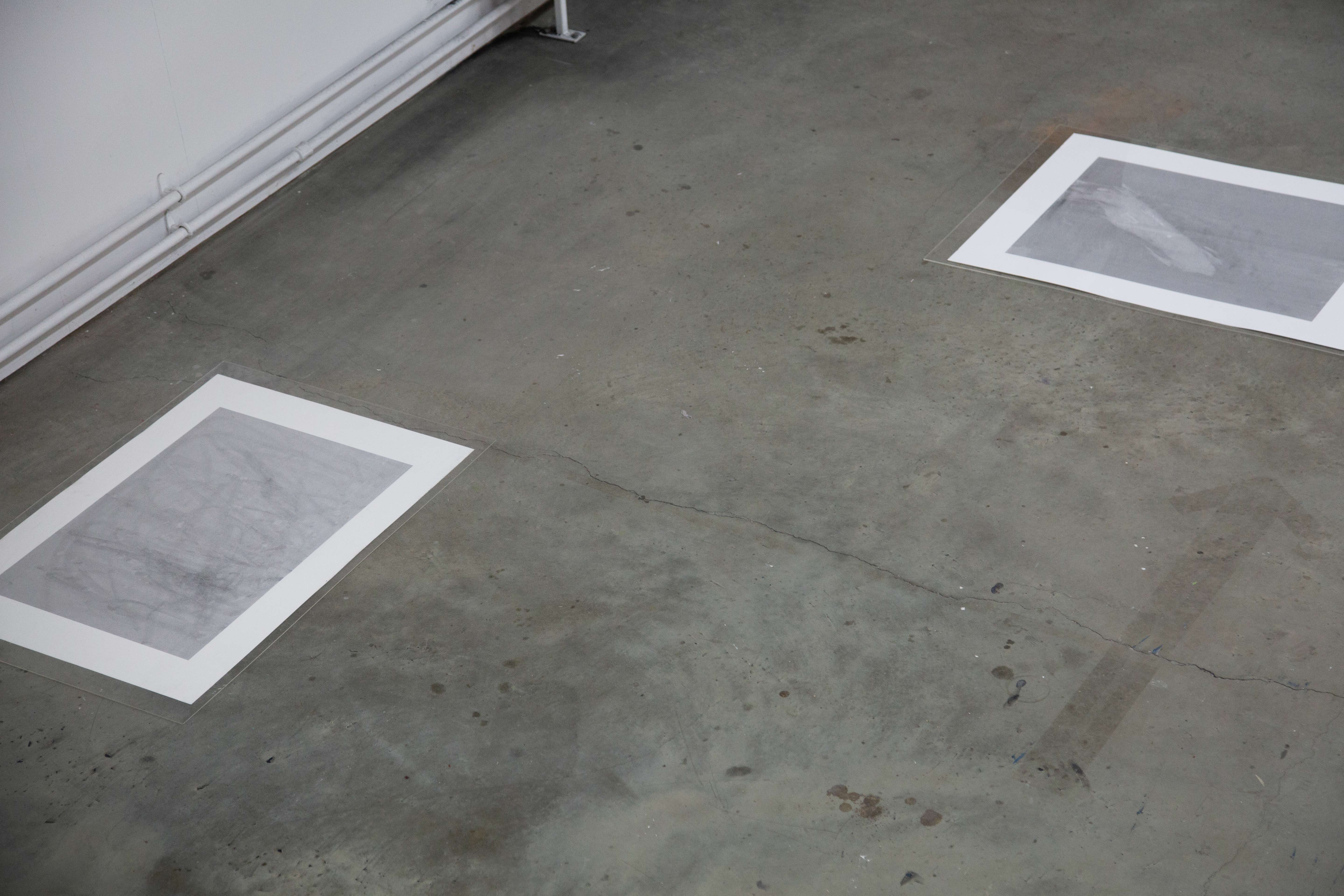 A photograph of two grey abstract prints with white boarders, spaced apart on a textured concrete floor, in perpendicular relation to one another.