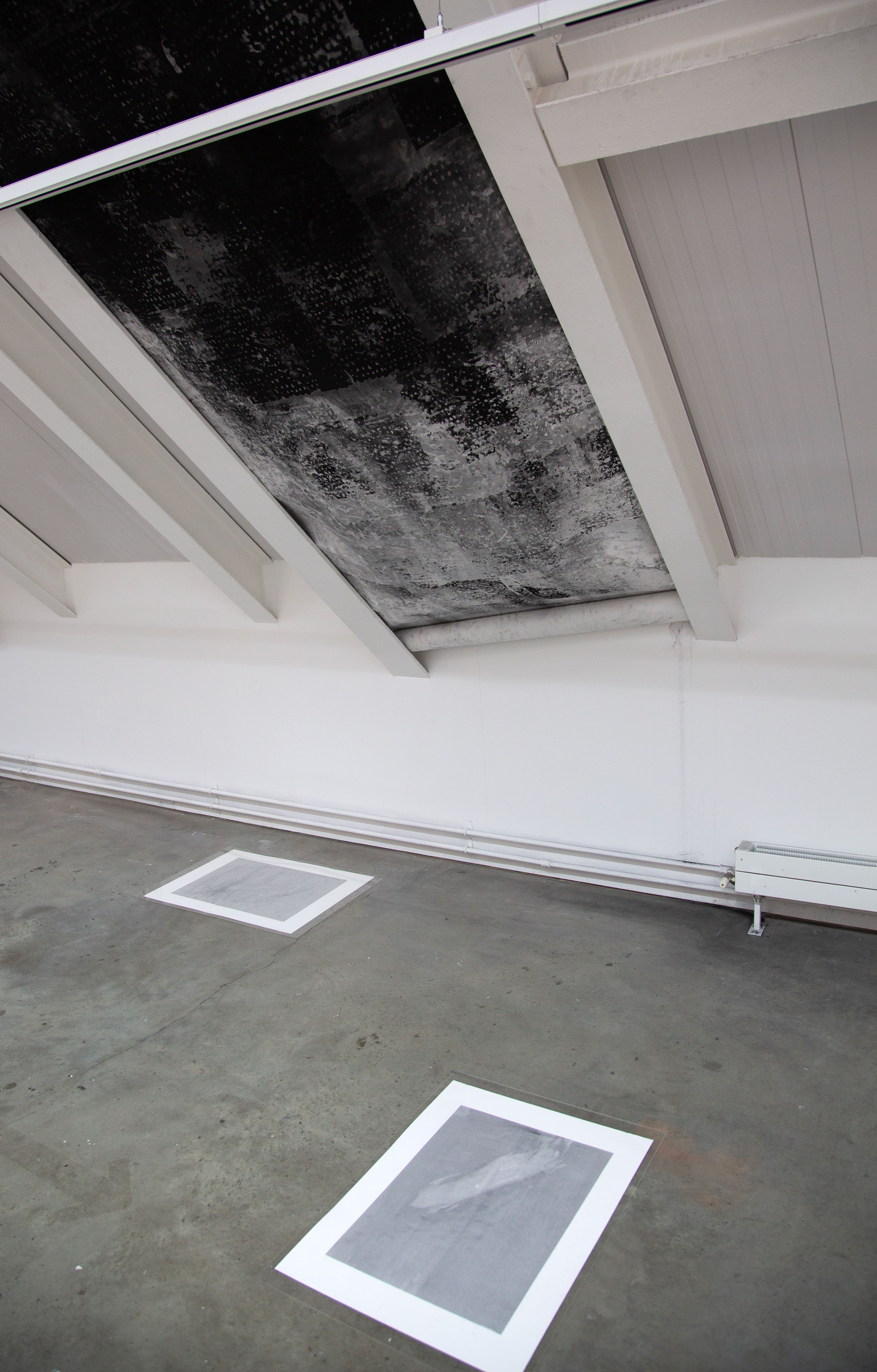 A photograph of a large black and white abstract charcoal drawing installed on an angled wall in between white mettle beans, with two smaller light grey prints on the concrete floor beneath it.