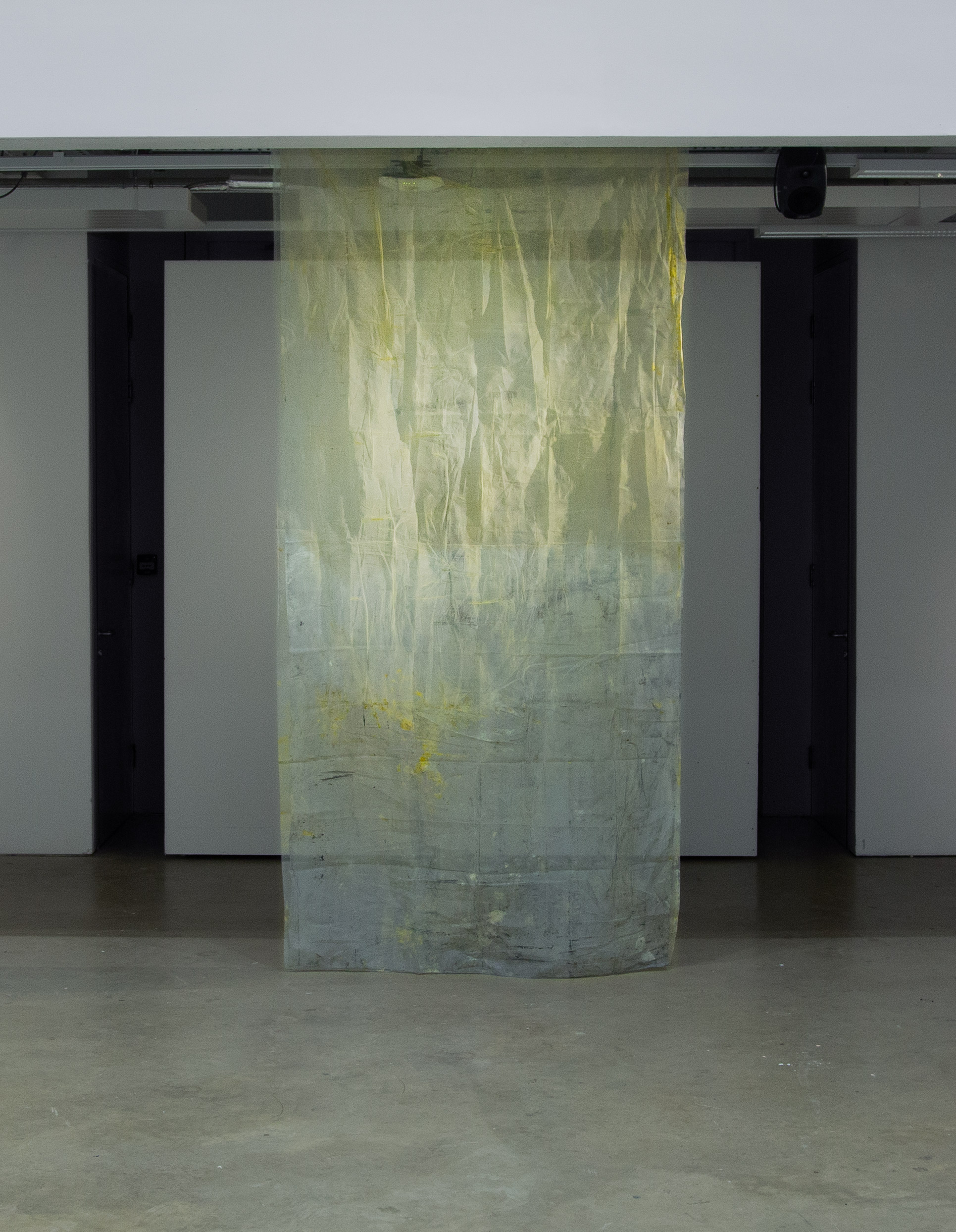 A vertical, translucent, yellow fabric, catching light, cascades from the galleries white-grey ceiling and rests lightly on the room's concrete floor.