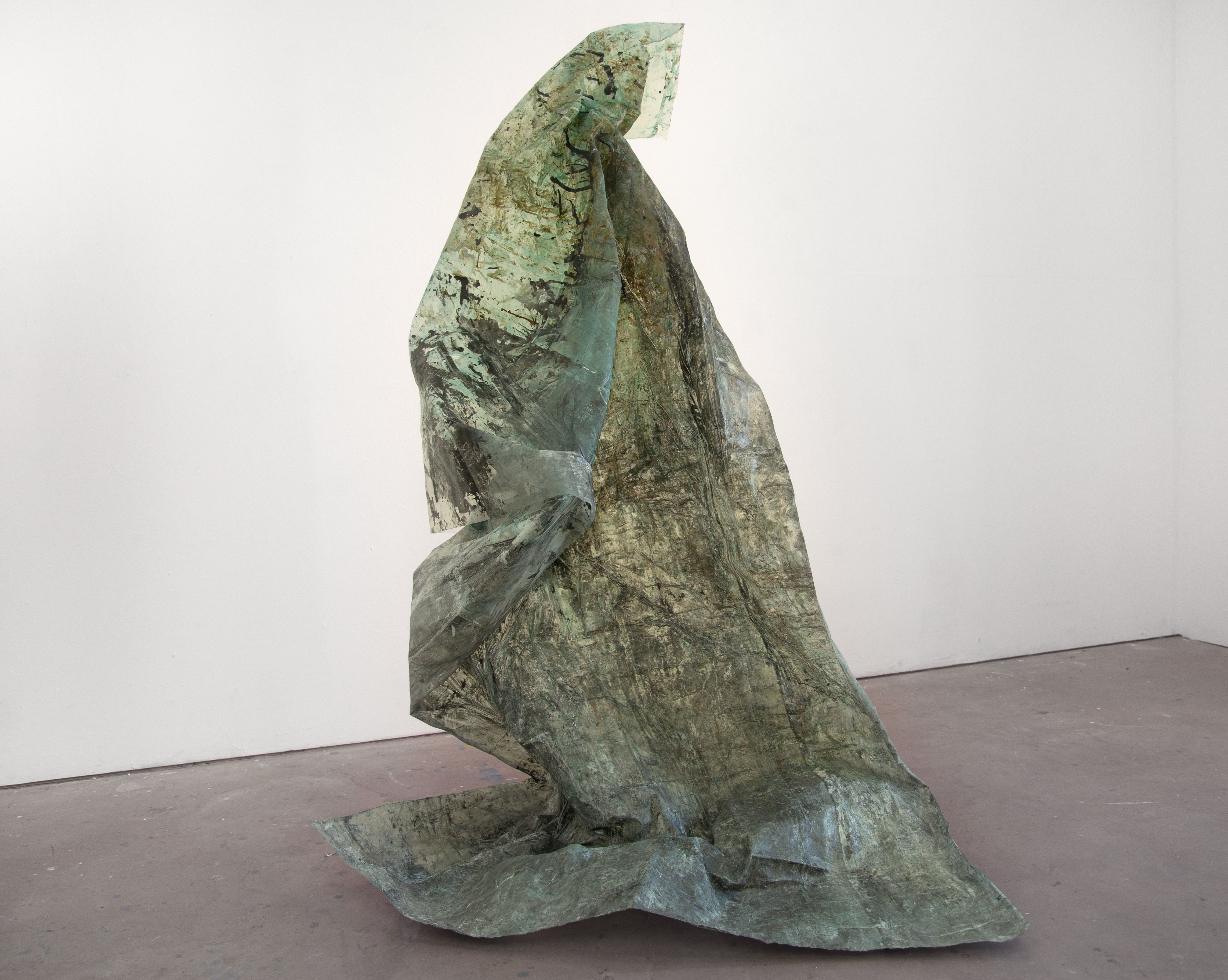 A large, vertical, green, textural sculpture, standing on a light grey gallery floor with a white wall behind it, made out of a translucent fabric material, shiny in some sections.