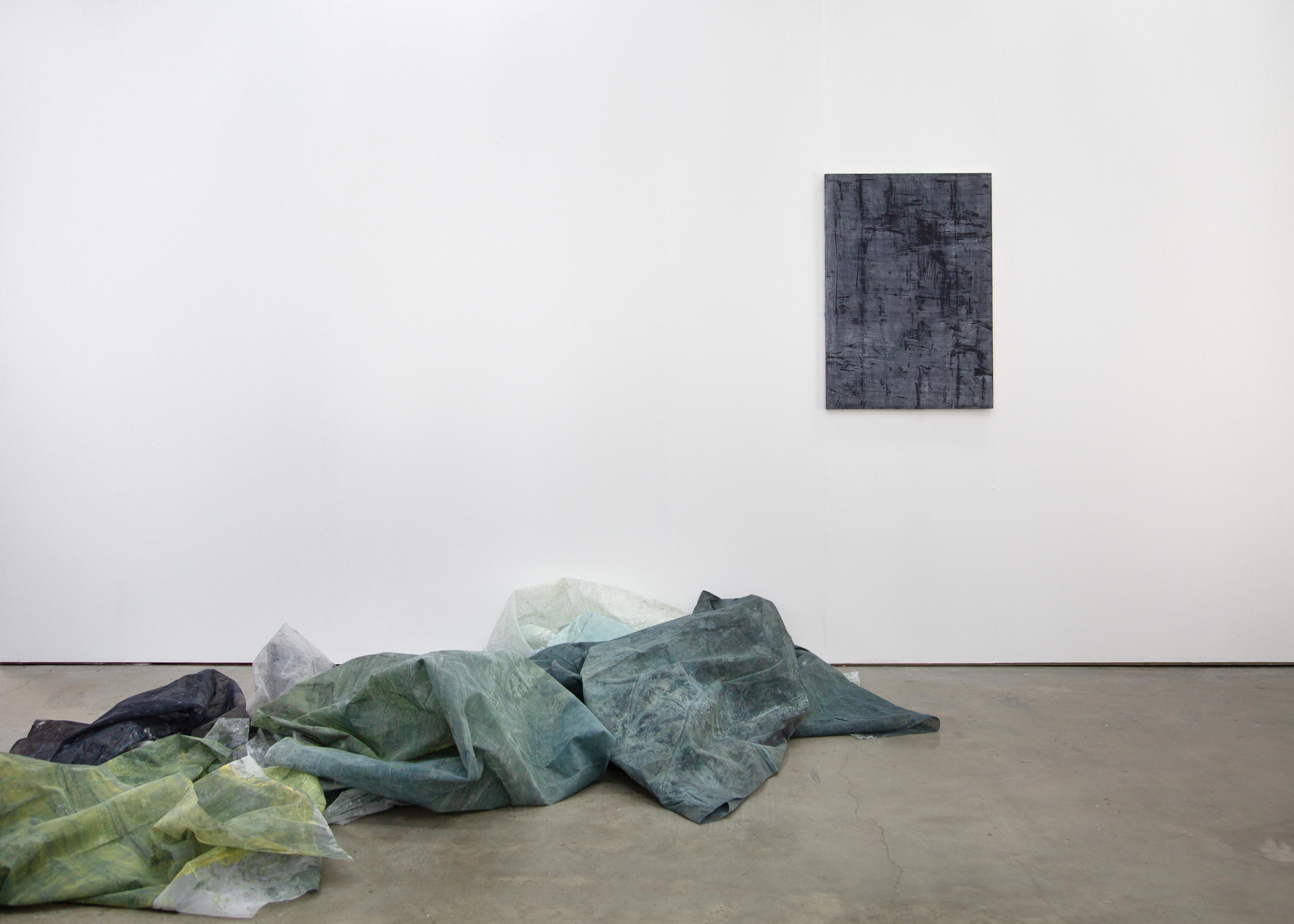 Photograph of a white gallery wall with a blue abstract painting, by artist Caroline Coolidge, hung vertically in the center right of the composition, and an artfully arranged pile of blue and green painted mesh fabrics on the floor, corresponding to the painting.
