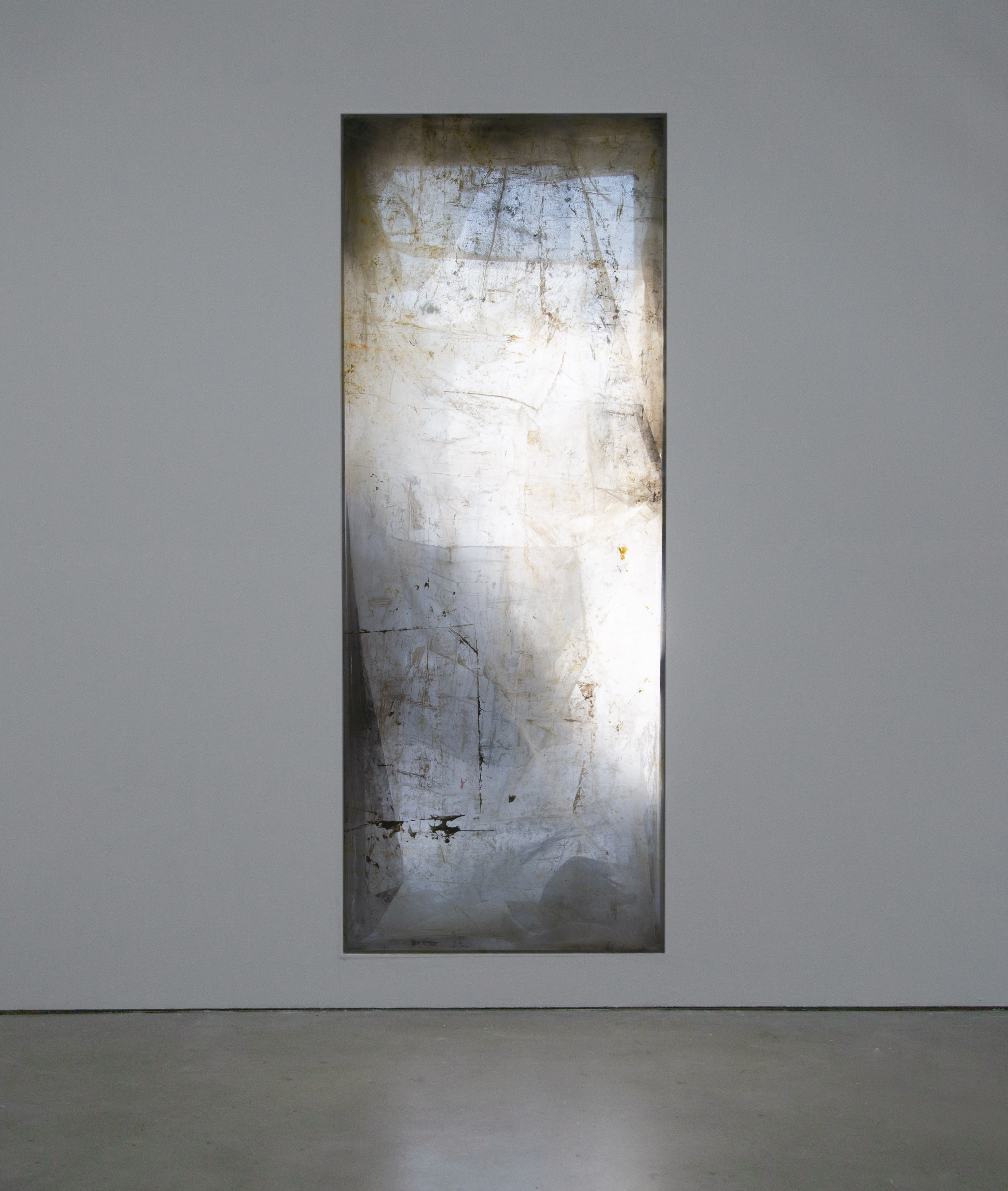 A light-emanating, vertically tall, pastel, and textured artwork by Caroline Coolidge embedded in a white wall that is lit from the space behind it.