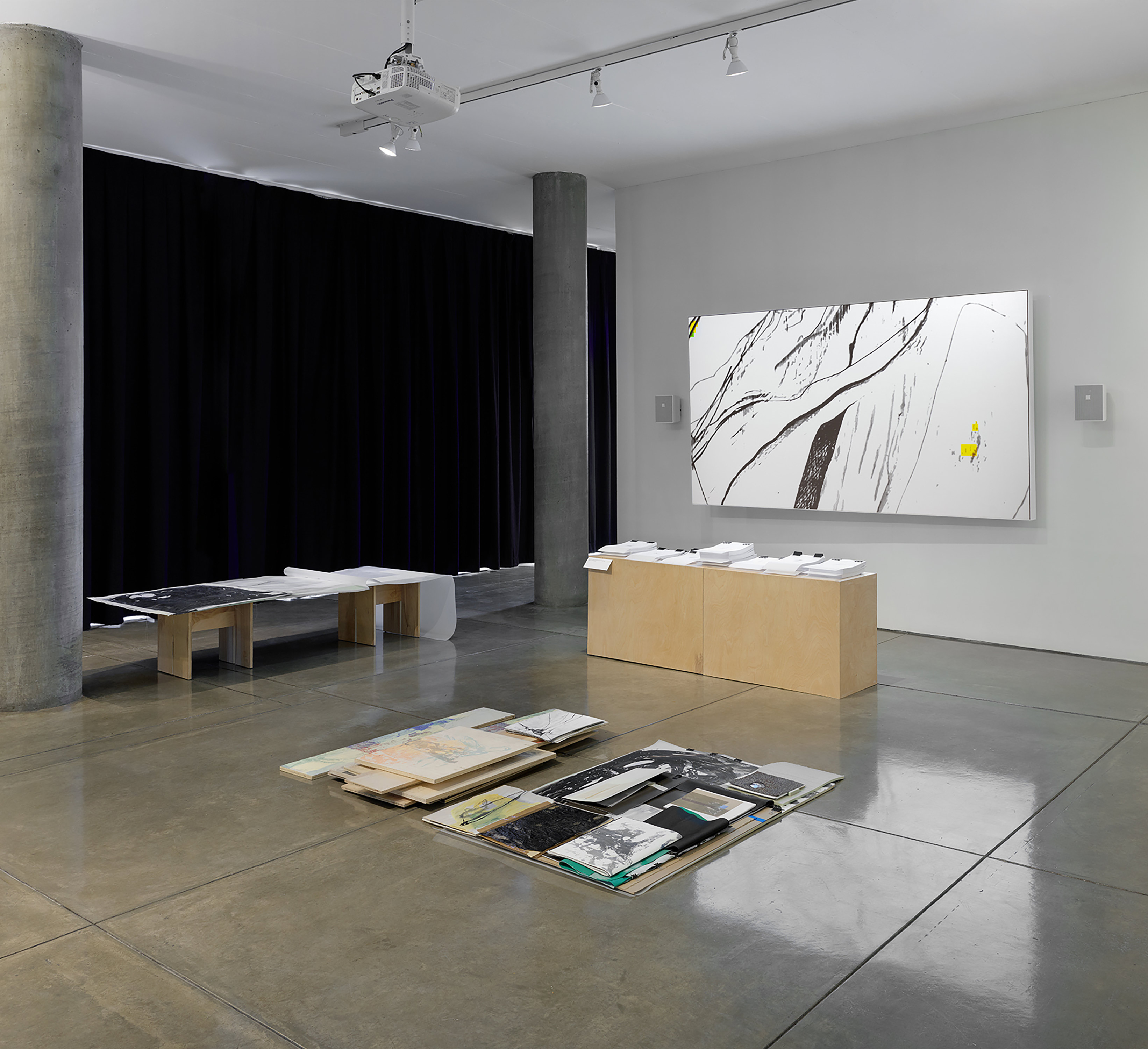 A gallery space with an art installation by Caroline Coolidge comprised of a projected image on a screen mounted to the far right wall, a table with stacks of paper on it, abstract paintings on wood and paper stacked on the shiny concrete floor, and a large abstract book form resting on a medium-high table to the back-left of the works on the floor.