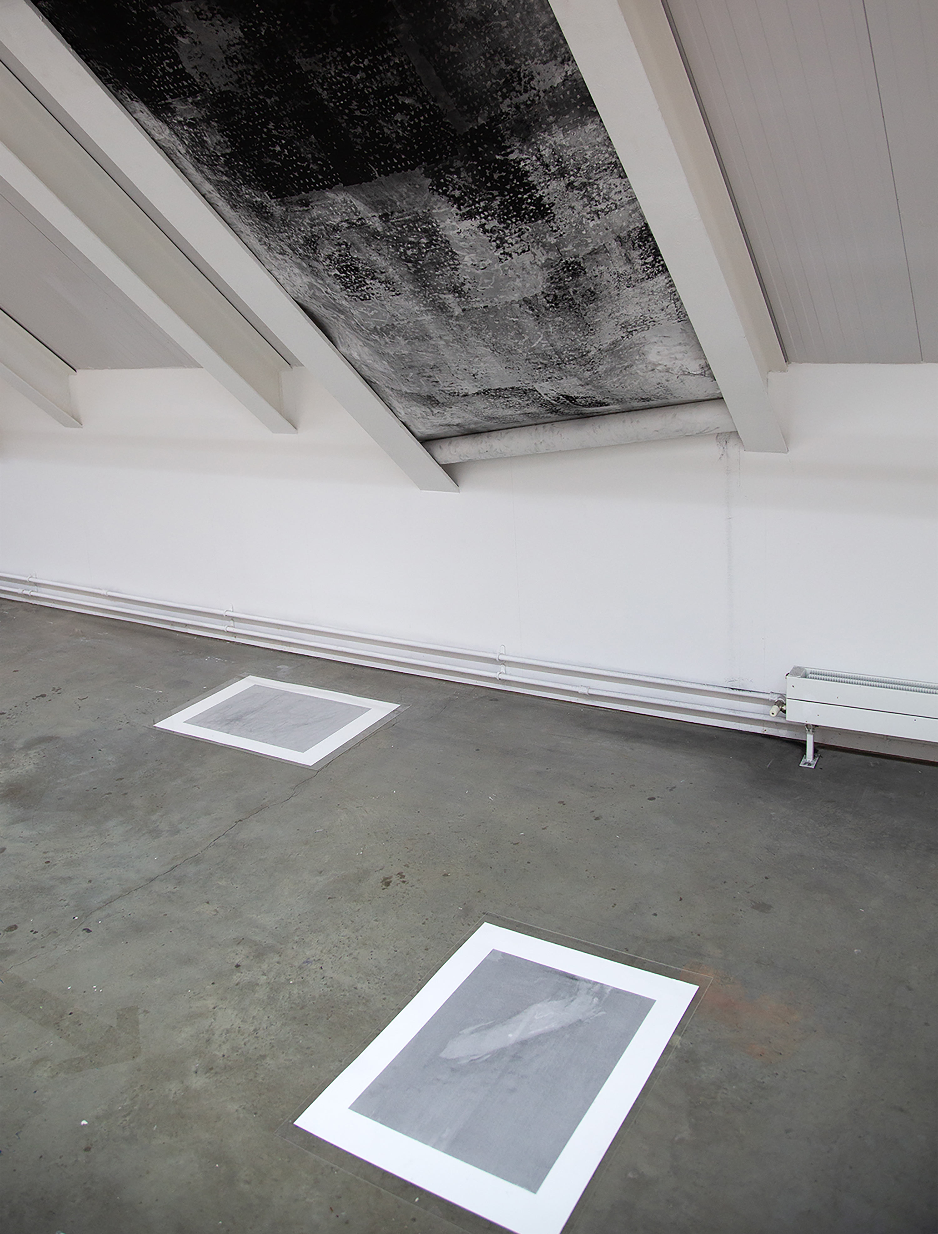 A photograph of a large, black and white, abstract, highly textural, charcoal drawing, installed between two white slanted metal beams and rolled up at the bottom, seen from an angled view, with the top of the drawing cut off, with two smaller light grey prints on the concrete floor beneath it. Art installation by Caroline Coolidge.