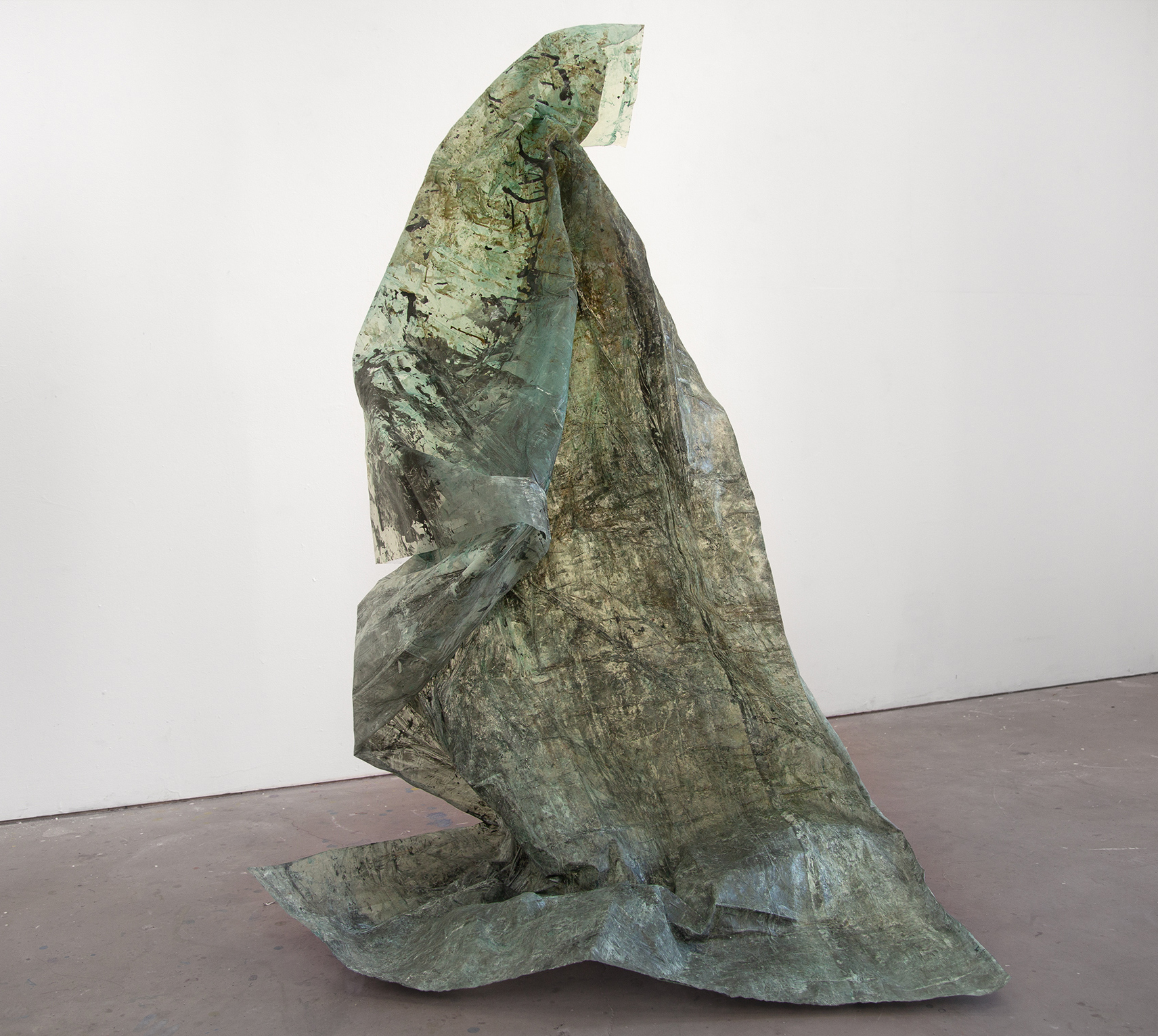 A large, vertical, green, textural sculpture by Caroline Coolidge, resting on a light grey gallery floor with a white wall behind it, made out of a translucent fabric material, shiny in some sections.