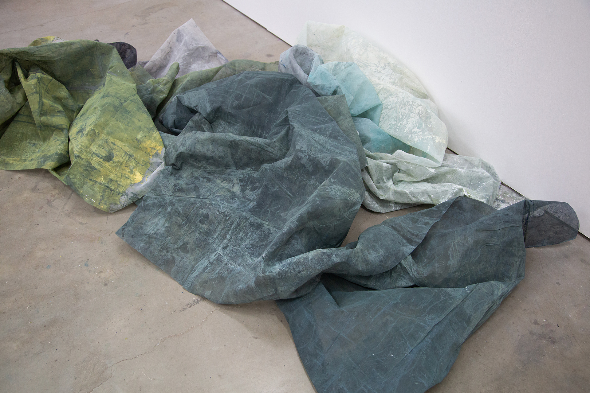 Photograph showing a closely cropped view of overlapping, bunched up mesh screens on a light-grey floor in four distinct shades of light and dark, blue and green. Artwork by Caroline Coolidge.