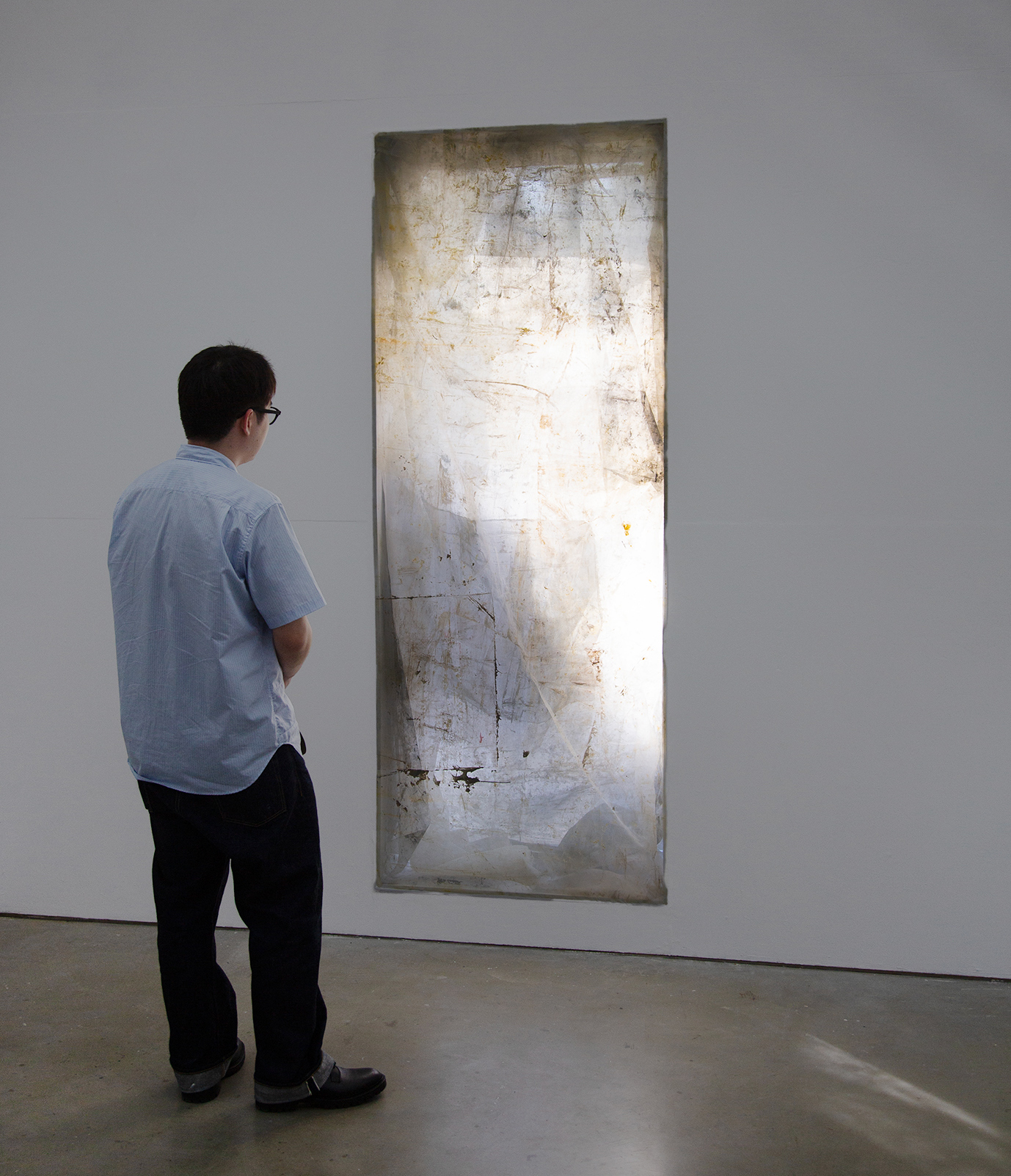 A long, vertical window covered in translucent layers of painted mesh screens, giving the appearance of a glowing impressionist painting. It is being contemplated by a dark-haired man in a blue shirt who is facing the artwork by Caroline Coolidge.