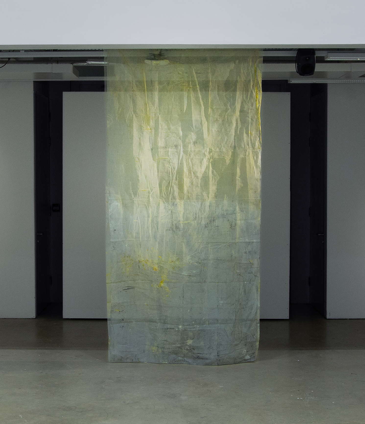 A vertical, translucent, yellow fabric, catching light, cascades from the gallery's white-grey ceiling and rests lightly on the room’s concrete floor, artwork created by Caroline Coolidge.