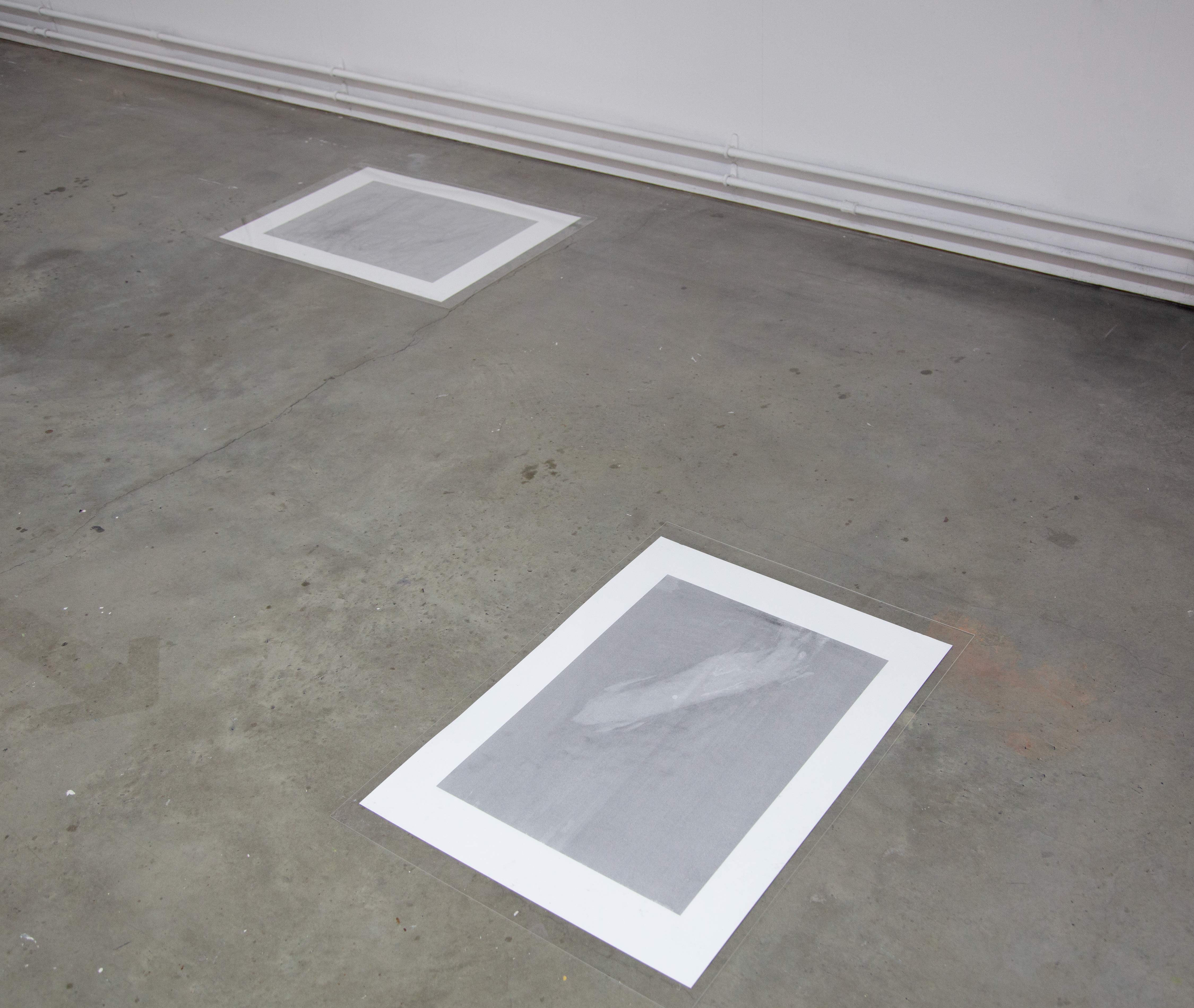 Two gray, monotone prints on paper by artist Caroline Coolidge lie on a gray concrete floor, not touching each other.