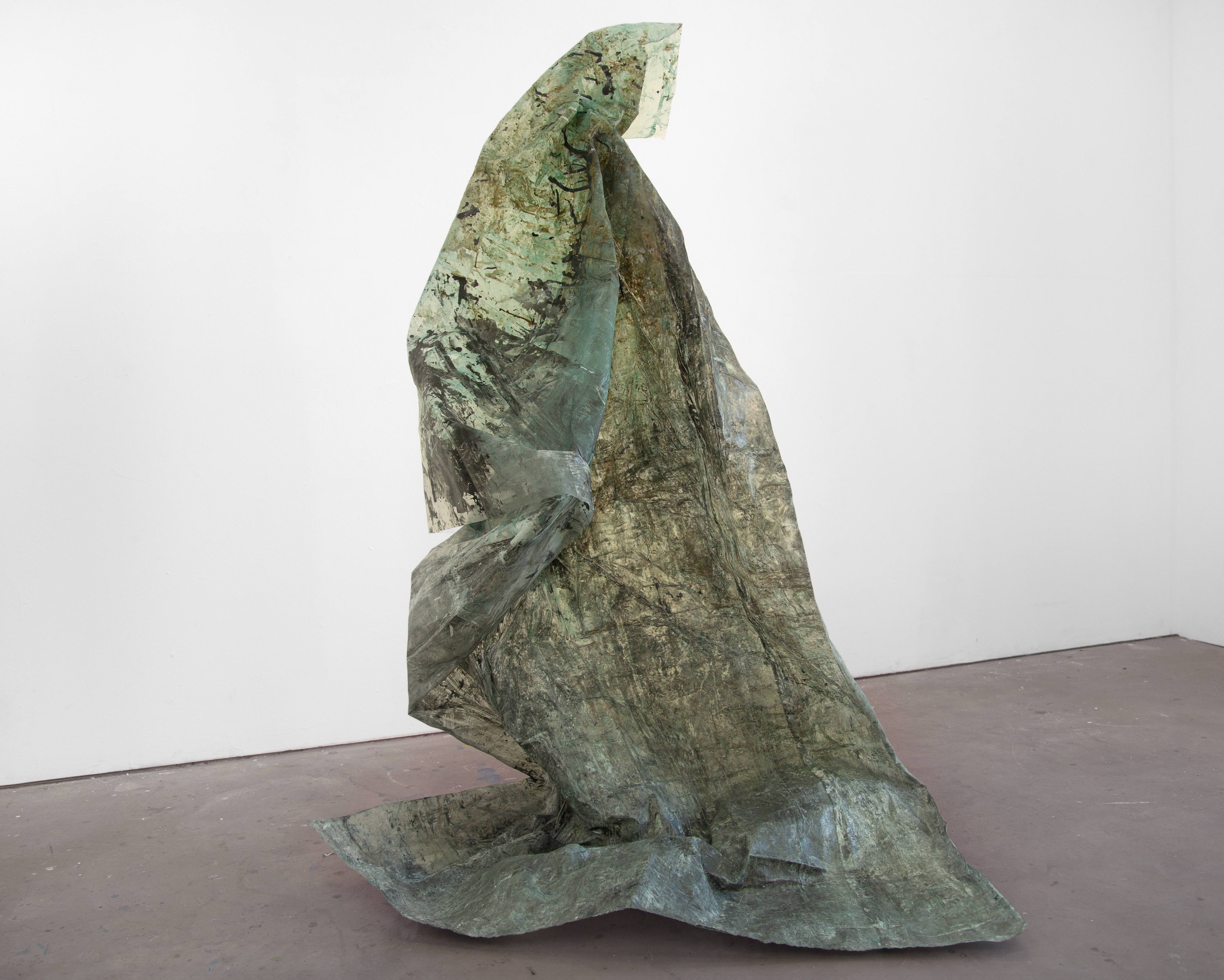 A large, vertical, green, textural sculpture by Caroline Coolidge, resting on a light grey gallery floor with a white wall behind it, made out of a translucent fabric material, shiny in some sections.